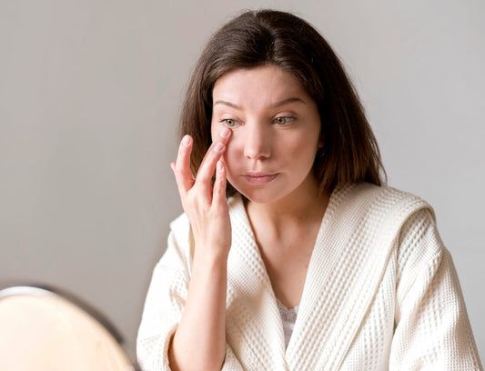 Woman in a robe touching the under-eye area while checking her skin in a mirror